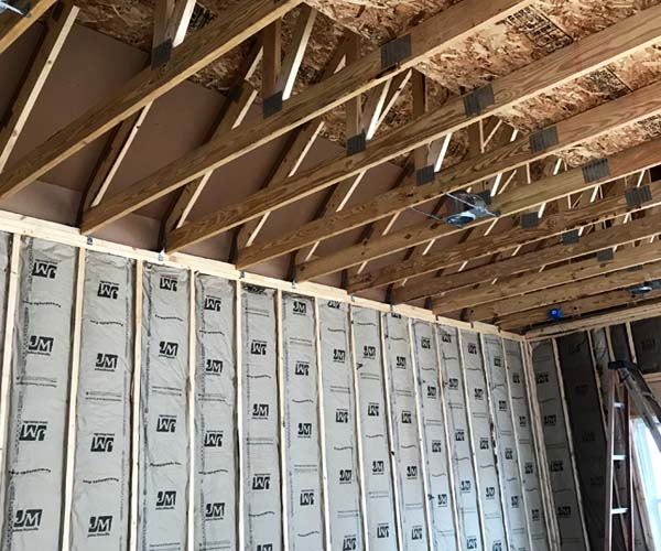 Fiberglass batt insulation installed between wall studs in a residential home under construction, with exposed wooden ceiling trusses and OSB roof decking visible.