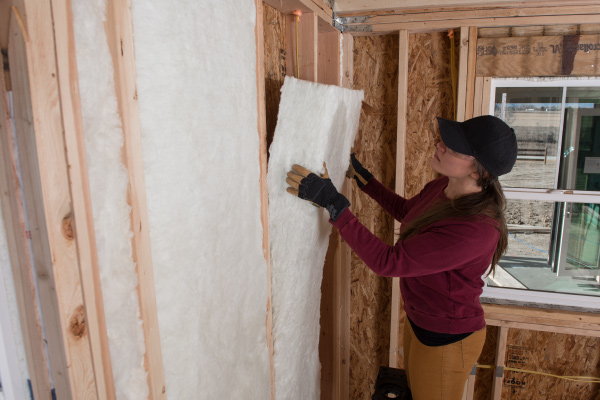 Worker installing white fiberglass batt insulation in a wall cavity.