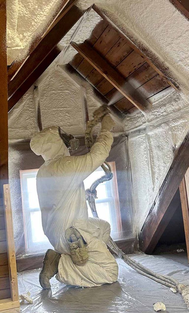 Spray foam technician in protective suit applying insulation to attic ceiling.