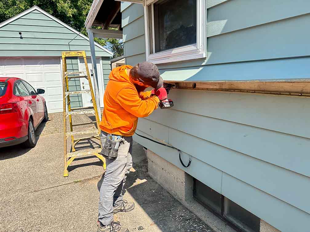 Technician drilling exterior siding to create access points for cellulose insulation installation.