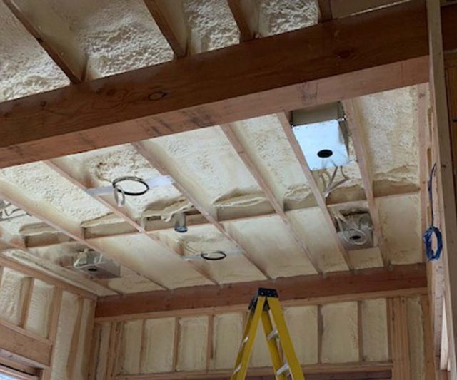 Spray foam insulation applied between floor joist bays with recessed light housings and a ladder visible below.