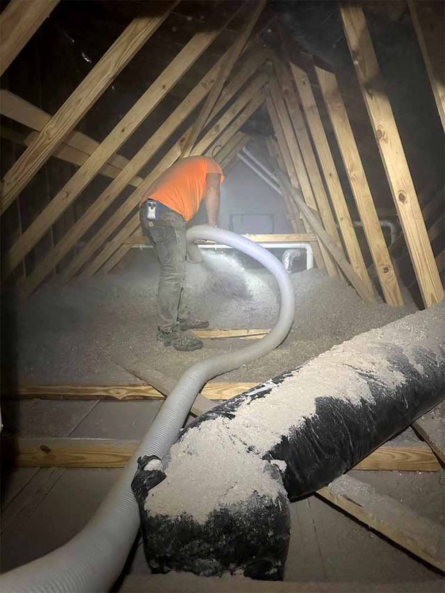Whitson Insulation installer blowing cellulose insulation across an attic floor under wood roof framing, with the hose running past insulated flex ductwork in Grand Rapids, Michigan.